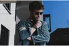 A man wearing sunglasses and a corduroy jacket leans against a wall, looking down. Produktbild-Vorschau