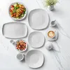 A collection of white ceramic tableware, including plates, bowls, a mug, and a creamer, arranged on a marble surface. A salad with grilled chicken is also present. Voir les images du produit