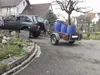 A dark gray SUV is parked next to a trailer carrying three blue barrels. The scene appears to be a residential area with houses in the background. Produktbild-Vorschau