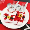 Christmas-themed cutlery holders shaped like Santa's suit are placed on a white plate, which is on a red placemat. Cutlery is inserted into the holders. Produktbild-Vorschau
