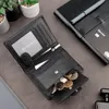 A black leather wallet is open, showing its contents, including coins and cards. It is placed on a dark surface next to a notebook and a pen. In the background, there are small potted plants. Produktbild-Vorschau