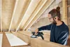 A man is sanding wooden beams in an attic space. Voir les images du produit