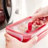 Hands are shown closing a red food container filled with pasta, with a microwave in the background. Produktbild-Vorschau