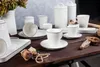 A collection of white ceramic tableware with dark rims is arranged on a wooden table, with some dried decorative elements and flowers in the background. Anteprima dell’immagine prodotto