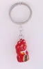 A red Maneki-neko (lucky cat) charm with a bell on a keychain, set against a white background. Produktbild-Vorschau