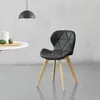 A black upholstered chair with wooden legs is placed on a gray floor in front of a textured white wall. A white table with two green fruits and a white vase is to the left of the chair. A black pendant lamp hangs from the ceiling above the table. Produktbild-Vorschau