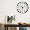 A wall clock is mounted on a white wall above a dining table with a bowl of fruit and a framed picture. Voir les images du produit