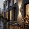 A modern house exterior at dusk with a wall-mounted light illuminating the entrance and fallen leaves on the wet ground. Podgląd zdjęcia produktu