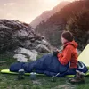 A person is sitting in a sleeping bag next to a tent, holding a mug, with mountains in the background. Produktbild-Vorschau