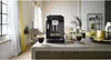 A black DeLonghi coffee machine is shown on a kitchen counter, with two cups of coffee in front of it. The background includes a window with curtains, a chair, and shelves with decorative items. Produktbild-Vorschau