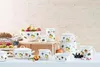 A collection of fruit-themed cookware and serving dishes arranged on a wooden crate and a white surface, with a light wooden plank background. Produktbild-Vorschau