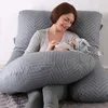 A woman is holding a baby while sitting on a bed with a large J-shaped pillow. Náhled obrázku produktu