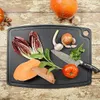 A black cutting board with various vegetables and a knife arranged on it, set on a rustic wooden table. Anteprima dell’immagine prodotto