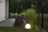 A garden scene at dusk with a glowing spherical lamp illuminating a large ornamental grass and lavender bushes. Anteprima dell’immagine prodotto