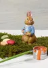 A ceramic bunny figurine dressed in a blue jacket, holding a palette and paintbrush, surrounded by moss and Easter eggs, with a can of orange paint and a paintbrush in the foreground. Podgląd zdjęcia produktu