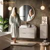 A woman in a bathrobe stands in a modern bathroom, looking into a large round mirror above a vanity. The vanity has a white countertop with a sink and a gold faucet, and white ribbed drawers with gold handles. There is a tall white ribbed cabinet to the ri Produktbild-Vorschau