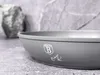 A close-up shot of a gray pot with a logo and signature on its side, placed on a textured gray surface with a blurred rock in the background. Podgląd zdjęcia produktu