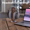 A pair of headphones rests on a wooden stand next to a laptop on a wooden table. Produktbild-Vorschau