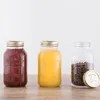 Three Kilner jars are arranged on a wooden surface against a white background. The leftmost jar contains a dark red substance, likely jam or sauce. The middle jar holds a bright yellow liquid, possibly juice. The rightmost jar is empty and contains coffee  Podgląd zdjęcia produktu