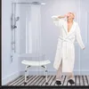 An elderly man in a bathrobe stands in a shower stall, yawning, with a shower stool in the foreground. Produktbild-Vorschau