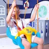 A young boy is happily swinging indoors on a blue and yellow swing set, with a nautical-themed room in the background. Produktbild-Vorschau
