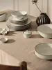 A collection of ceramic tableware, including plates, bowls, and a small lidded dish, arranged on a textured beige tablecloth. A dark, ribbed vase with a dried sprig of greenery is also present. Produktbild-Vorschau