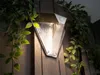 A solar-powered outdoor light with a motion sensor is mounted on a wooden fence, surrounded by green foliage. Produktbild-Vorschau