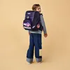 A young girl wearing a large purple backpack with star patterns and colorful patches on her jeans, looking over her shoulder. Produktbild-Vorschau