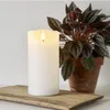 A white LED candle sits on a wooden surface next to a potted plant. Produktbild-Vorschau