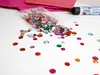 A collection of colorful rhinestones spilled from a plastic bag onto a white surface, with a tube of glue in the background. Anteprima dell’immagine prodotto