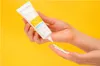 A person's hands are shown squeezing sunscreen from a tube onto their palm against a bright yellow background. Náhled obrázku produktu