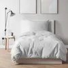 A white duvet and pillow are neatly arranged on a bed frame, with a wall-mounted lamp and a small side table with books and a mug to the left. Two abstract art pieces hang on the wall behind the bed. Produktbild-Vorschau