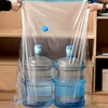 Two large water bottles are placed inside a large clear plastic vacuum storage bag, which is being held open by two hands. The bag is positioned in front of a wooden shelf unit. Voir les images du produit