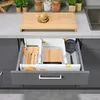 A drawer organizer filled with kitchen utensils and a small cutting board is pulled open, revealing its contents. Produktbild-Vorschau