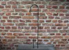 A black metal shepherd's hook garden stake stands upright against a brick wall, with its base resting on a wooden surface. Produktbild-Vorschau