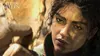 Close-up of a woman's face with dirt on her skin, looking intensely forward. The background is blurred and appears to be a rocky, desert-like environment. Náhled obrázku produktu