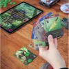 A hand holding game cards over a wooden table with a tablet and other game components. Anteprima dell’immagine prodotto