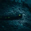 A black flashlight with green indicator lights lies on a dark, rocky surface. Produktbild-Vorschau