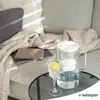 A glass water pitcher and a glass of water with lemon slices are placed on a round glass table in front of a sofa. Náhľad obrázku produktu