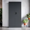 A dark gray metal cabinet with two doors and a lock is shown in a room with a concrete wall and floor. There are plants in pots on the right side of the cabinet. Produktbild-Vorschau
