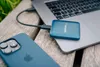 A blue Verbatim SnapBack SSD is connected to a laptop, with an iPhone in the foreground, all resting on a wooden surface. Náhľad obrázku produktu