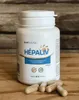 A white bottle of Hepaliv supplements with several capsules scattered in front of it, placed on a wooden surface with a textured wooden background. Produktbild-Vorschau