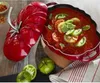 A red Staub cocotte shaped like a tomato, filled with tomato soup and garnished with tomato slices and basil, sits on a wooden table next to green tomatoes and a striped napkin. Náhľad obrázku produktu