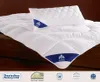 A white quilted duvet and pillow with blue trim and branding, laid on a wooden surface. Produktbild-Vorschau