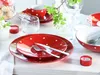 A table setting with red and white polka dot tableware, including plates, a cup, and a bowl, along with cutlery, a glass, and a small candle. A sprig of white flowers adds a decorative touch. Produktbild-Vorschau