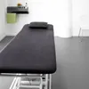A treatment table with a dark grey cover and a dark grey pillow, placed in a clean, modern room with a shelf holding a green towel, a flower, and bottles. Produktbild-Vorschau
