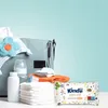 A collection of baby care items, including a diaper bag, baby powder, bottle, diapers, and a pack of baby wipes, arranged on a surface against a light blue background. Podgląd zdjęcia produktu