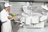 A person in a white uniform and cap is using a high-pressure washer to clean a large industrial machine covered in white foam. Náhled obrázku produktu