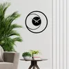 A modern black metal wall clock with a circular design is mounted on a white wall. A lush green plant is in the foreground to the left, and a small table with potted plants and books is in the foreground to the right. Anteprima dell’immagine prodotto