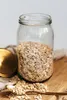 A glass jar filled with oats sits on a wooden board, with a golden lid and spoon nearby. Produktbild-Vorschau